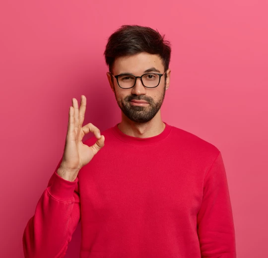 Homme barbu avec des lunettes, vêtu d’un pull rouge, faisant le geste OK avec la main devant un fond rose uni.Les gîtes du haut bois 2 Le Haut Bois, 25650 Ville-du-Pont, France